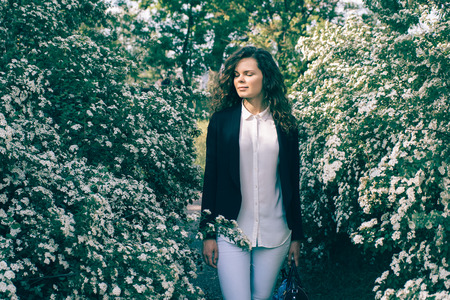Young beautiful woman in a jacket and jeans stands in a garden among white flowers