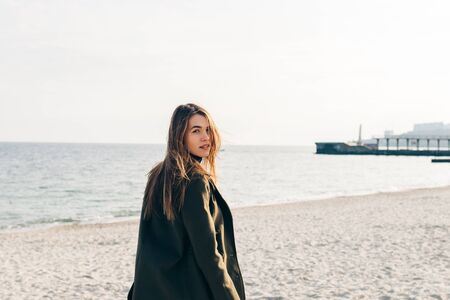 Young beautiful woman in a green coat with long hair walking along an empty beachの写真素材