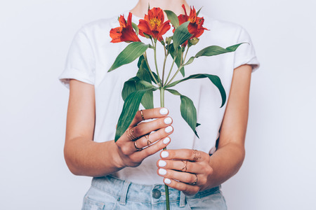 Cropped image of a girl in jeans and a T-shirt holding a red flower in her hands with a manicure on a white backgroundの写真素材