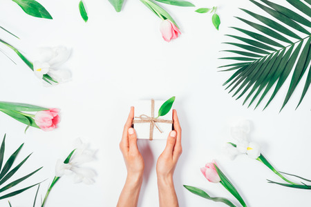 Female hands holding a small gift box among the flowers on a white background, top viewの写真素材