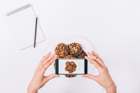 Female hands photographing on a mobile phone a plate with cookies on a white table top viewの写真素材