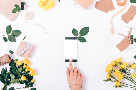 Mobile phone with blank screen in female hands and a festive composition of yellow flowers on a white table top viewの写真素材
