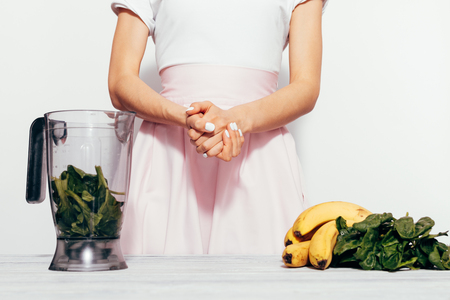 Young woman in a pink skirt prepares smoothies from a banana and spinach, close-upの写真素材
