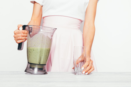 Slender young woman in a pink skirt pours a green smoothie from a banana and spinach into a glassの写真素材