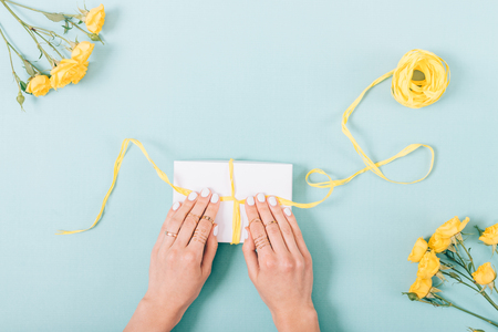 Woman packing a present. Top view of female hands with rings tying a bow on the gift box among yellow roses on trendy blue background. Flat lay composition.の写真素材