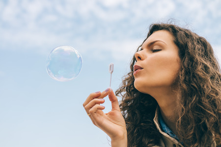 Portrait of young woman with brown curly hair blowing soap bubbles on blue sky background. Carefree...