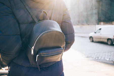 Rear view woman with leather backpack wearing down jacket goes on city street at morning or evening....