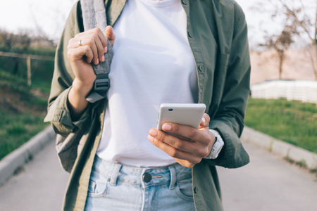 Close-up woman with backpack uses smart phone outdoors. Cropped photo of girl wearing casual outfit...