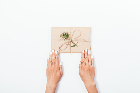 Women's hands with white manicure and gold rings hold a paper gift tied with twine and decorated with a pink flower, top view. Minimal flat lay composition with copy space.の写真素材