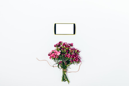 Top view summer composition of bouquet and smart phone with blank screen above it. Flat lay minimal floral arrangement of pink flowers and mobile device on white background.の写真素材