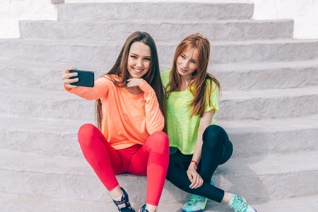 Two girl friends in sporty outfits do a joint selfie sitting on the steps outdoors. Both happy young females relaxing and smiling looking into a smart phone.の写真素材