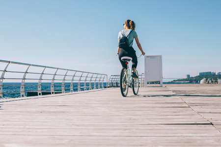 Beautiful young woman riding bicycle on the waterfront at sunny summer day. Back view female with a backpack looking at the sea while cycling.の写真素材