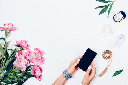 Women's hands holding smart phone on a white background among bouquet of peonies and feminine jewelry. Flat lay floral frame with empty place in the center.の写真素材