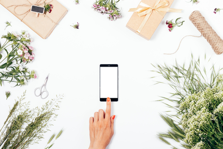 Female using smart phone among gifts and summer field flowers, top view. Flat lay festive composition woman is touching screen with her index finger.の写真素材