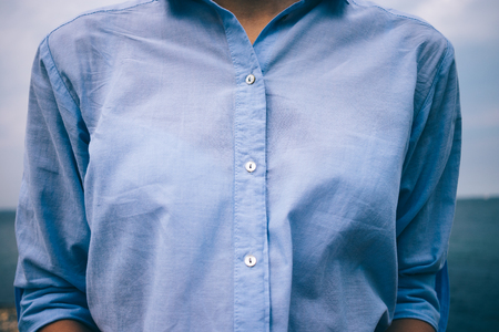 Close-up details of women's casual clothing. Unrecognizable young stylish woman dressed in blue cotton shirt with rolled up three-quarters sleeves on sea background.の写真素材