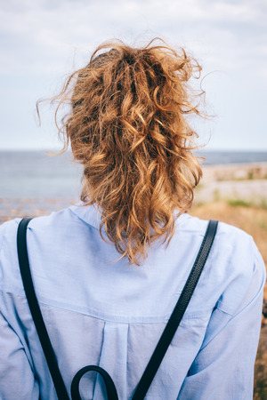 Rear view of young woman with thick curly hair in ponytail sitting on the seaside. Vertical photo of unrecognizable female wearing blue shirt and backpack relaxing and looking away.の写真素材