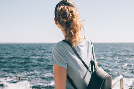 Candid image of young woman looking at the sea horizon, view from the back. Female with curly red hair wearing blue t-shirt and black backpack standing on the coast in the beautiful morning sunlight.の写真素材