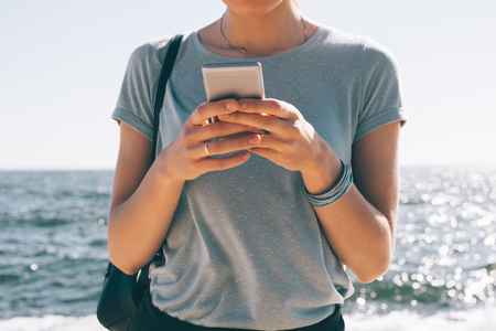 Fashionable young woman standing texting on the background of sea and sun flare. Cropped image of female wearing blue t-shirt and black shoulder bag using smart phone on summer day.の写真素材