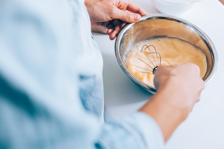 Candid image woman mixing batter for pancakes, close-up. Female's hands holding bowl whisking milk and eggs on white table.の写真素材