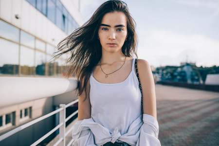 Beautiful young woman wearing white top posing at city street at summer day. Girl with fluttering...