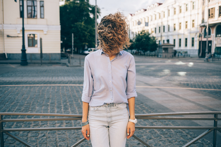 Candid lifestyle photo of fashionable female wearing blue shirt and jeans posing in old city at sunny summer day. Young woman standing on street with curly hair blown by the wind, covering her face.の写真素材