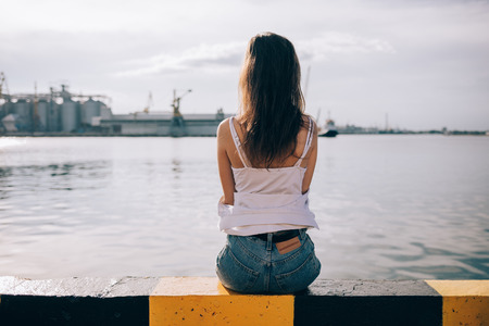 Rear view young woman with long hair sitting on parapet in the port looking at water. Girl wearing white top and blue jeans resting near the sea at sunny day.の写真素材