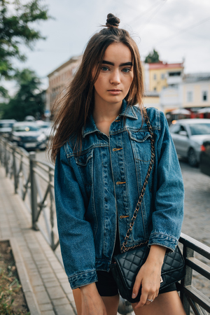 Young woman with long hair dressed in denim jacket standing on the street near road. Cute stylish teen girl posing in the city.の写真素材