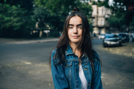 Teen girl dressed in blue jeans jacket looking at camera and smiling against the green trees and road. Portrait of happy beautiful young woman in the city on summer day.の写真素材