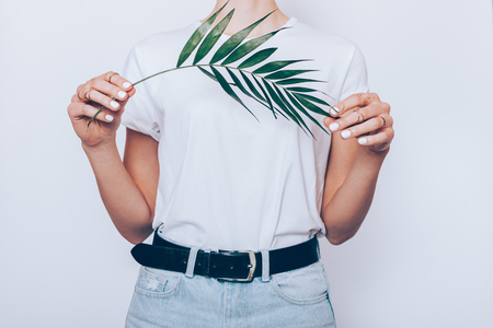 Slender young woman wearing blue high waist jeans with belt and plain t-shirt standing over white background holding green palm leaf, close-up.の写真素材