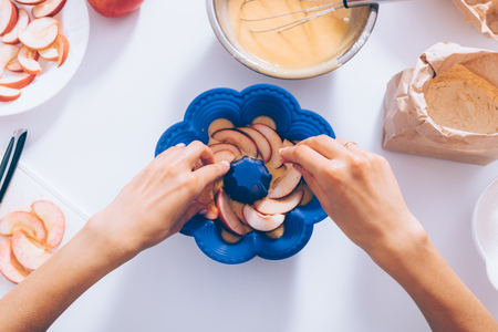 Female's hands add sliced fruits into baking dish for traditional apple pie near ingredients and kitchenware, top view. Woman cooking seasonal autumn cake, flat lay composition.の写真素材