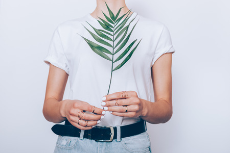 Close-up slender young woman wearing blue high waist jeans with black belt and plain t-shirt standing over white background holding green palm leaf.の写真素材