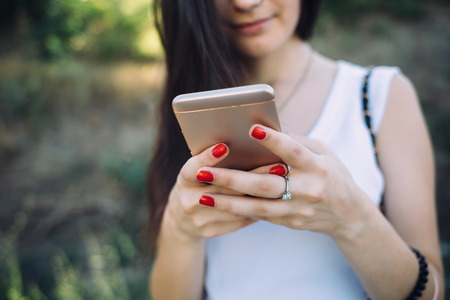 Happy young woman wearing white top using smart phone standing outdoors, close-up. Girl's hands holding mobile device on summer day at the background of green park grass.の写真素材