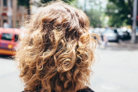 Woman's head with fluffy curly brown hair, back view. Close-up unrecognizable girl goes away at sunny day in the city.の写真素材