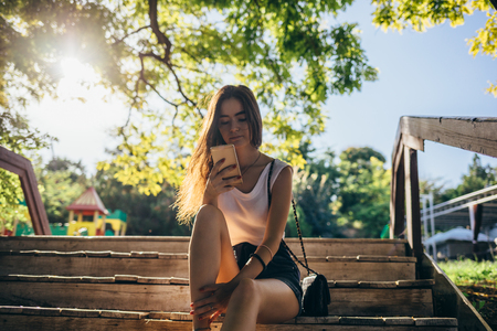 Young woman wearing shorts and white top is resting and using smart device, sun shining through the trees. Teen girl sitting on wooden bench outside in a green park looking at mobile phone.の写真素材