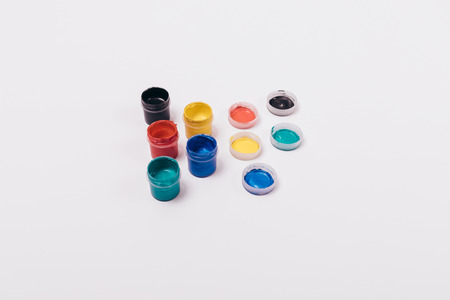 Group of multi-colored paints in open jars next to lids on solid white background, top view.