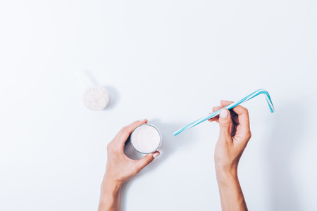 Woman making protein shake, top view. Female's hand holding glass of fresh made sports drink and blue straw near scoop with fitness supplement powder, flat layout on white background.の写真素材