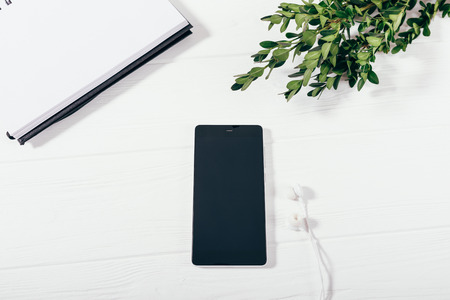 Close-up of mobile phone with blank screen next to headphones on white desk, top view. Flat lay composition of workplace with green plant, notepad and smart device in the center.の写真素材