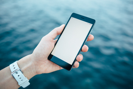Clean mobile phone screen template in female hand on blue water background at dusk. Girl holding smart device with blank display at sea, close-up.の写真素材