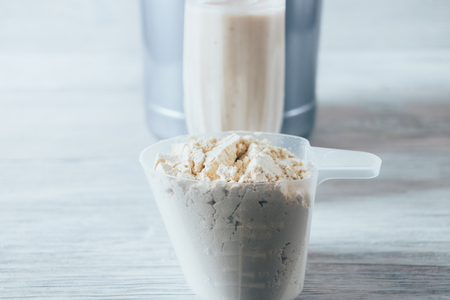 Close-up scoop with serving of dry protein powder and glass of sports nutrition drink on white wooden table. Portion of additive for muscle growth.の写真素材