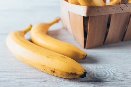 Close-up organic bananas near basket on white wooden table. Authentic image of yellow ripe tropical fruits with little dark spots.の写真素材