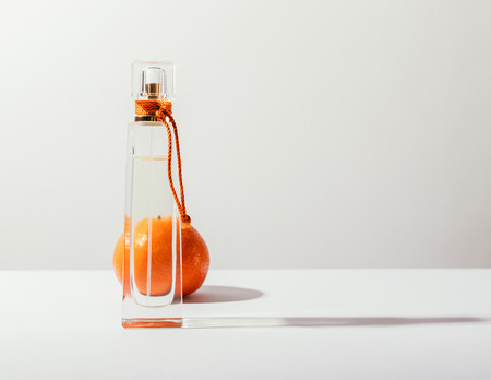 Citrus perfume in clear bottle and whole orange mandarin on the back. Toilet water with refreshing fruity smell standing on white background, casting shadow. Image of cosmetic product with copy spaceの写真素材