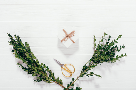 Gift box tied with twine next to scissors and two green branches of boxwood on white wooden table, top view. Flat layout of Christmas preparations, present wrapping and decorations.の写真素材