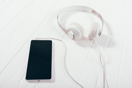 Headphones with mobile phone on white wooden desk, top view. Close-up composition with smart device connected to earphones to listen music, podcast or audiobook.の写真素材