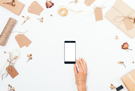 Top view female hand holding smart phone with blank screen. Romantic flat lay arrangement of dried roses, gifts, ribbons and candles on white background.の写真素材