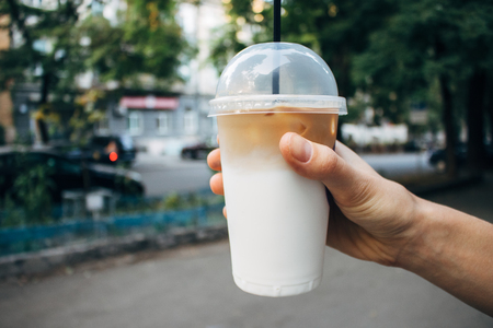 Hand holding coffee latte in plastic transparent cup with straw. Cold caffeine refreshment on background of city street at summer day.の写真素材