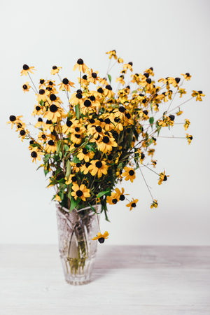 Beautiful bundle of small yellow flowers of rudbeckia with black middles. Vertical image lush simple rural eco bouquet in vintage crystal vase on white background.の写真素材