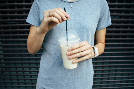Young woman holding coffee latte in plastic transparent cup with straw. Close-up female wearing grey t-shirt and jeans drinking cold caffeine refreshment standing outdoors.の写真素材