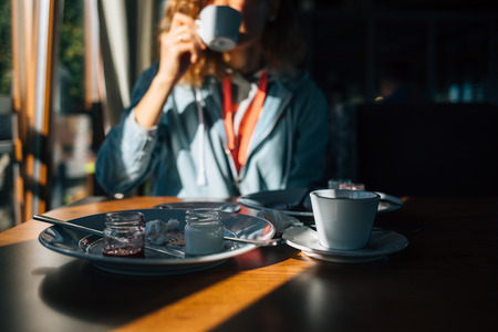 Selective focus remains of dessert on table and girl drinking coffee on background. Young woman indoors finished eating cake and now enjoying hot drink in cafe.の写真素材
