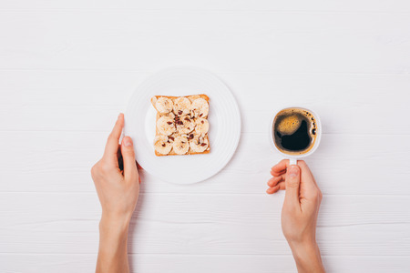 Healthy breakfast of sweet toast with banana, peanut butter and hazelnuts, flat lay on white wooden table. Woman's hands holding cup of coffee and plate with fruit sandwich with nuts and seeds, top view.の写真素材