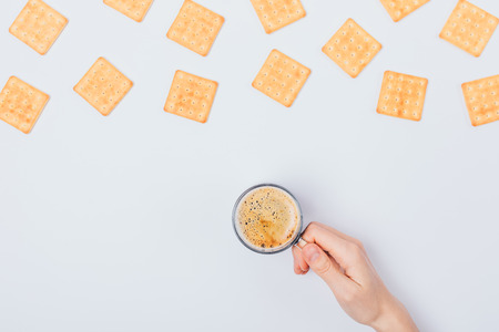 Female's hand holding cup of fresh coffee with creamy foam next to flat lay pattern frame of crackers on white background, top view.の写真素材